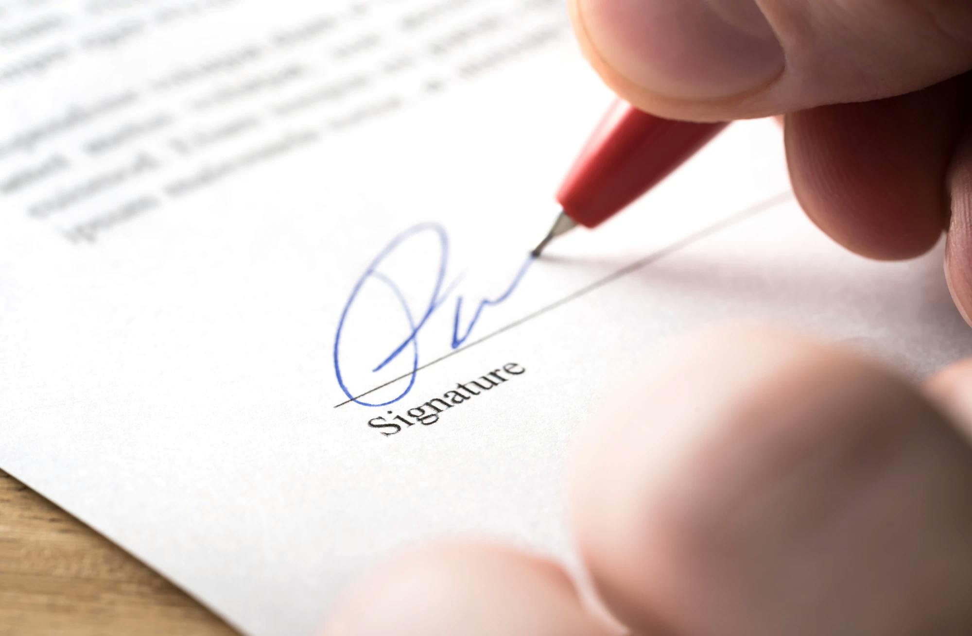 Close-up of a person signing a contract with a blue, fine-tipped pen, ostensibly a document related to the transfer of business.
