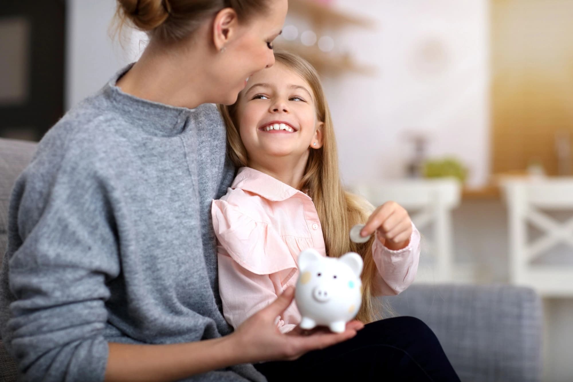 Young girl sitting on her moms lap putting money in a piggy bank